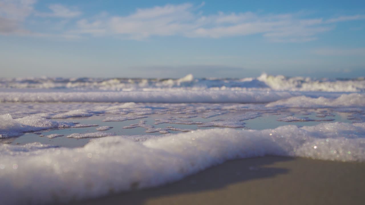 Small waves making foam on a beach. Low angle shot of blue water waves.