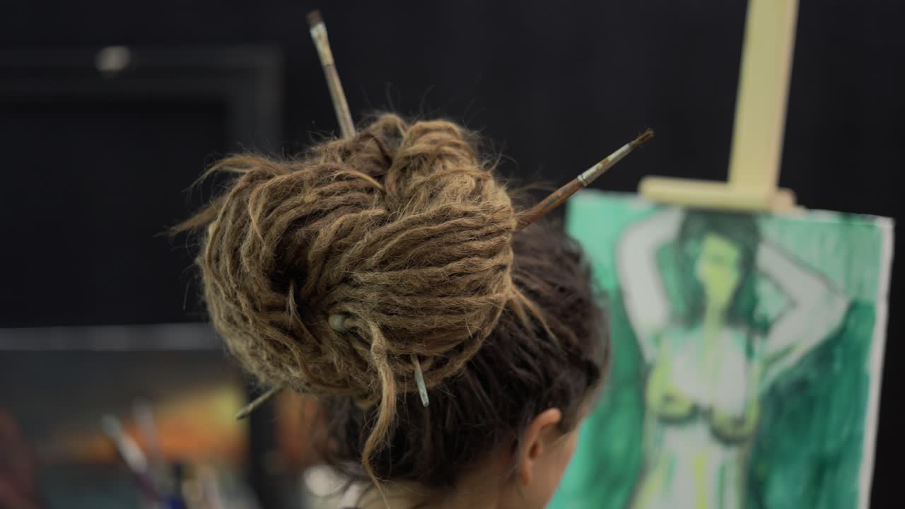 vista posterior de una pintora femenina con dreadlocks fijados por pinceles pintando en lienzo en un caballete