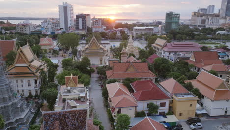 aerial video of phnom penh floats over temple rooftops at sunrise, their orange tiles glowing beneath the city skyline