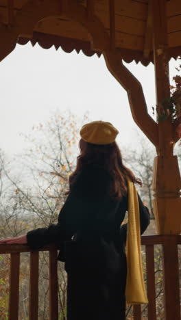 elegante dama en boina amarilla y silenciador explora un mirador de madera, haciendo una pausa para colocar su mano en la barandilla, mirando pensativamente en medio del telón de fondo de follaje de otoño