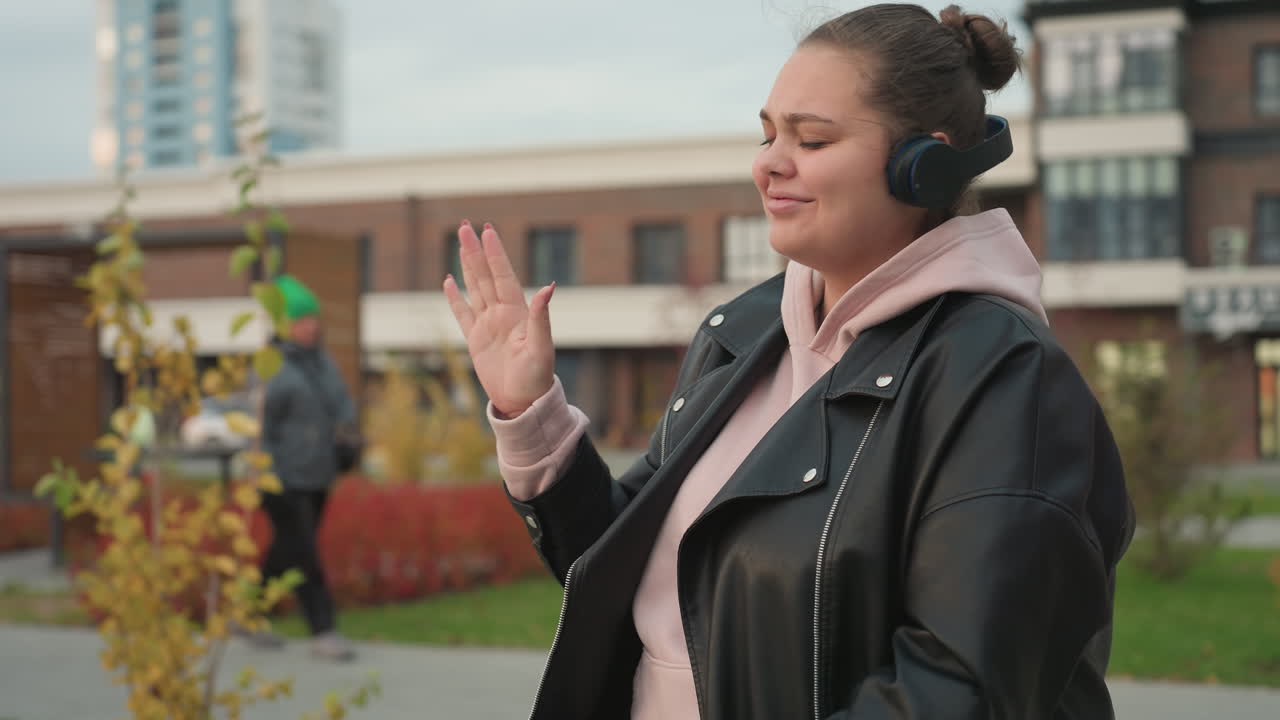 Happy woman wearing black leather jacket and pink hoodie dances joyfully while enjoying music outdoors as cyclist rides by and person walks nearby with autumn leaves swaying gently in breeze