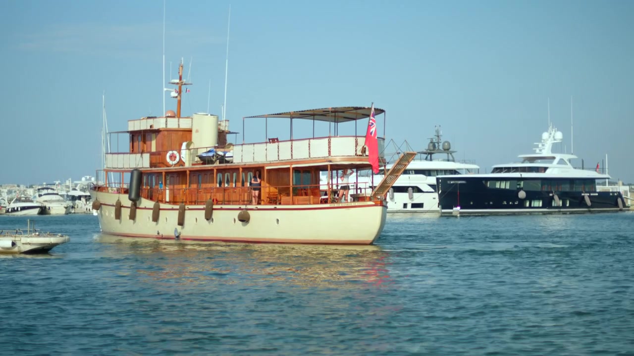 Sakin liman sularında seyreden, kırmızı bayraklı klasik ahşap yat, güneşli bir günde büyük modern lüks teknelerin yanından geçiyor.