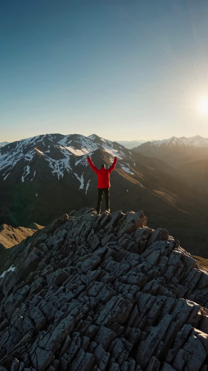 Un video aereo riprende una persona con una giacca rossa che si erge trionfante sulla cima di una montagna rocciosa