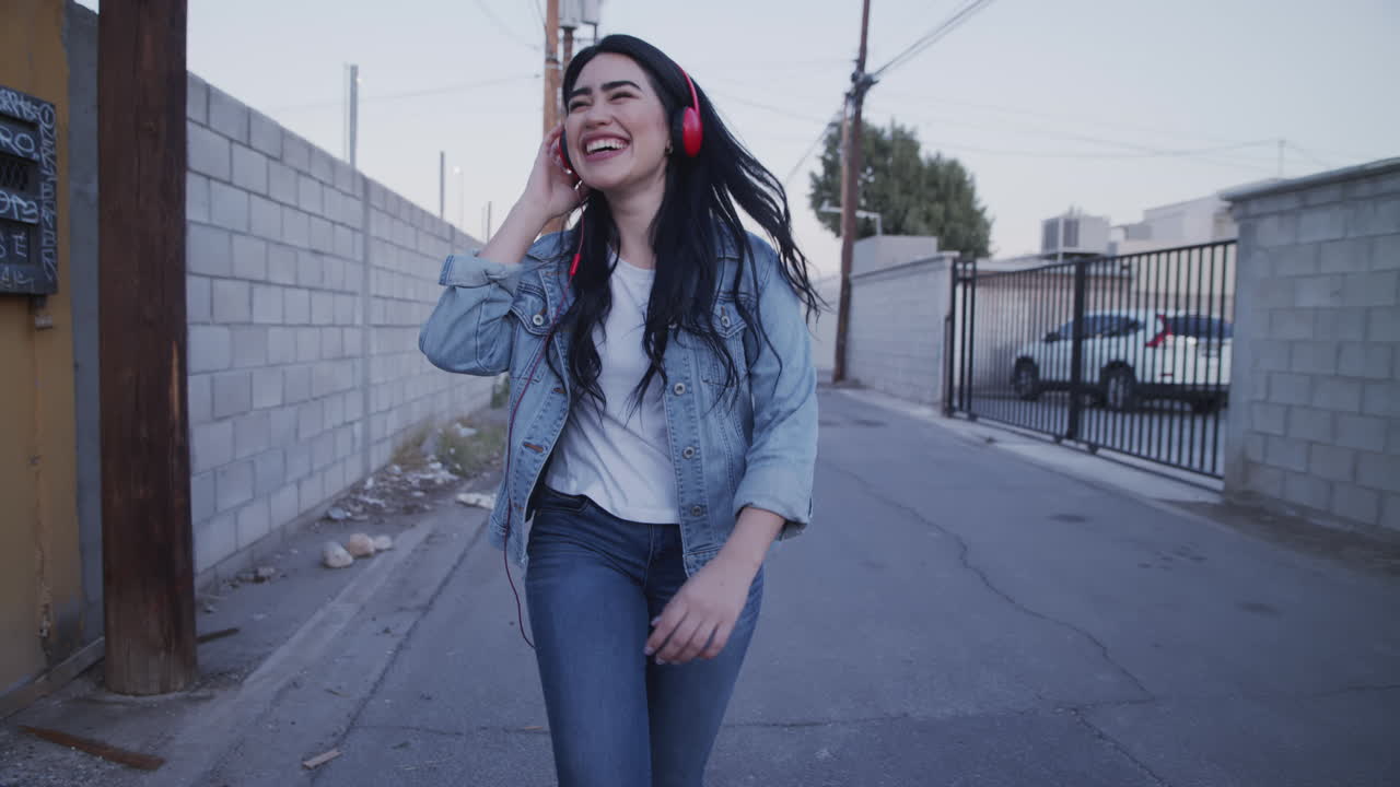 mujeres jóvenes felices sonriendo mientras escucha música y camina por el callejón, usando auriculares rojos