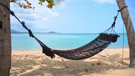 Empty-cottom-rope-net-hammok-with-spreader-bars-lashed-between-two-palm-trees-on-the-sandy-beach-in-front-of-the-turquoise-sea-at-tropical-island