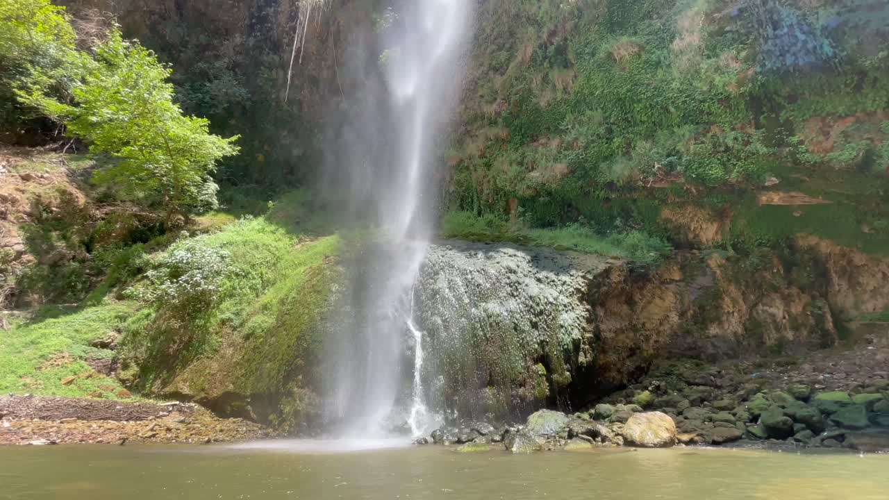 Water flows gracefully from a high cliff into a calm pool below, surrounded by vibrant vegetation and rocky terrain. Nature showcases its beauty in this serene landscape