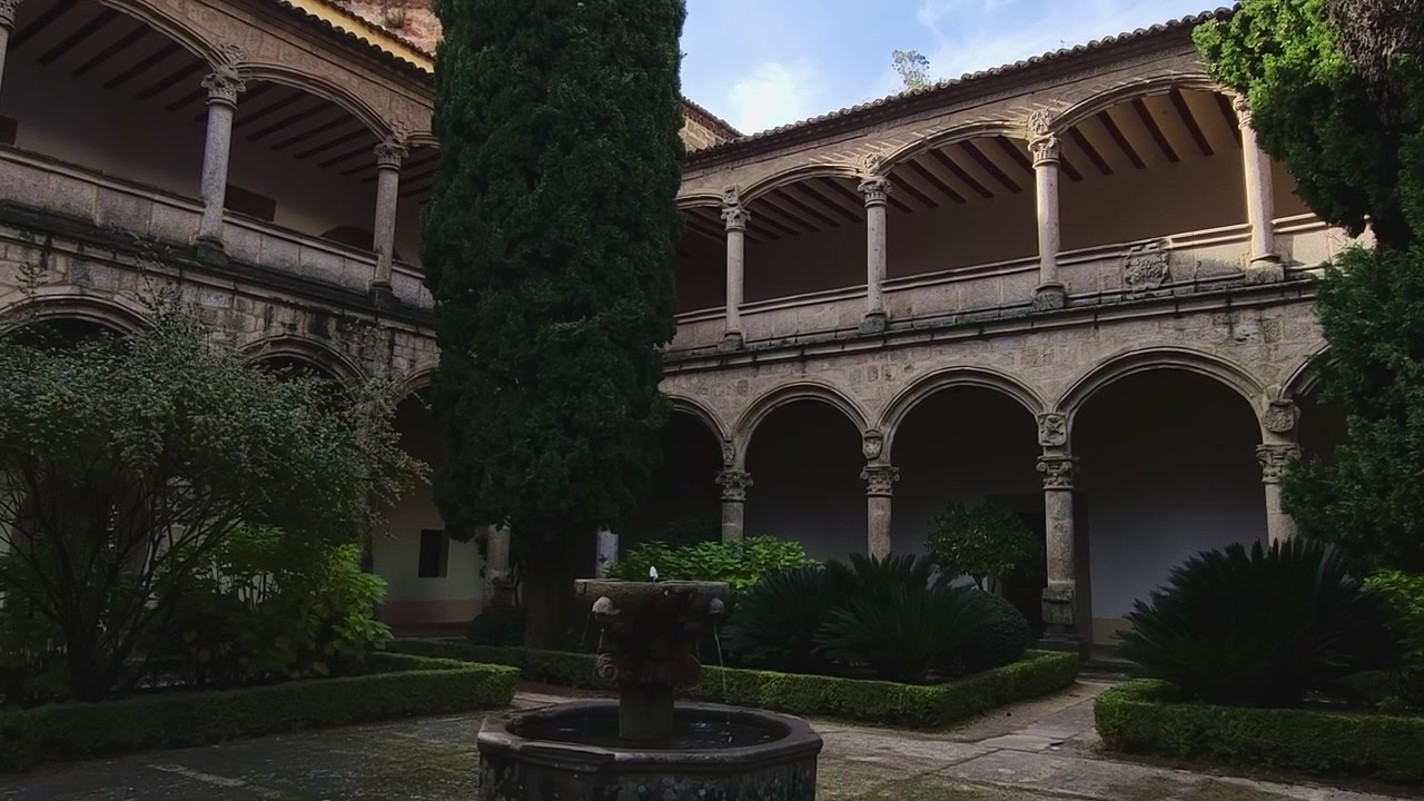 Cloître du monastère de Yuste. Arches en pierre sur deux niveaux, une fontaine en pierre et un grand cyprès pointant vers le ciel bleu. Espagne