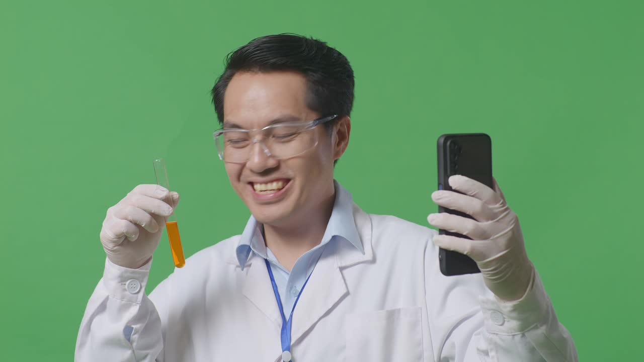 아시안 남성 과학자가 오렌지색 액체의 시험관을 들고 실험실의 녹색 화면 배경에 서있는 동안 스마트폰에서 비디오 통화를 하고 있습니다.