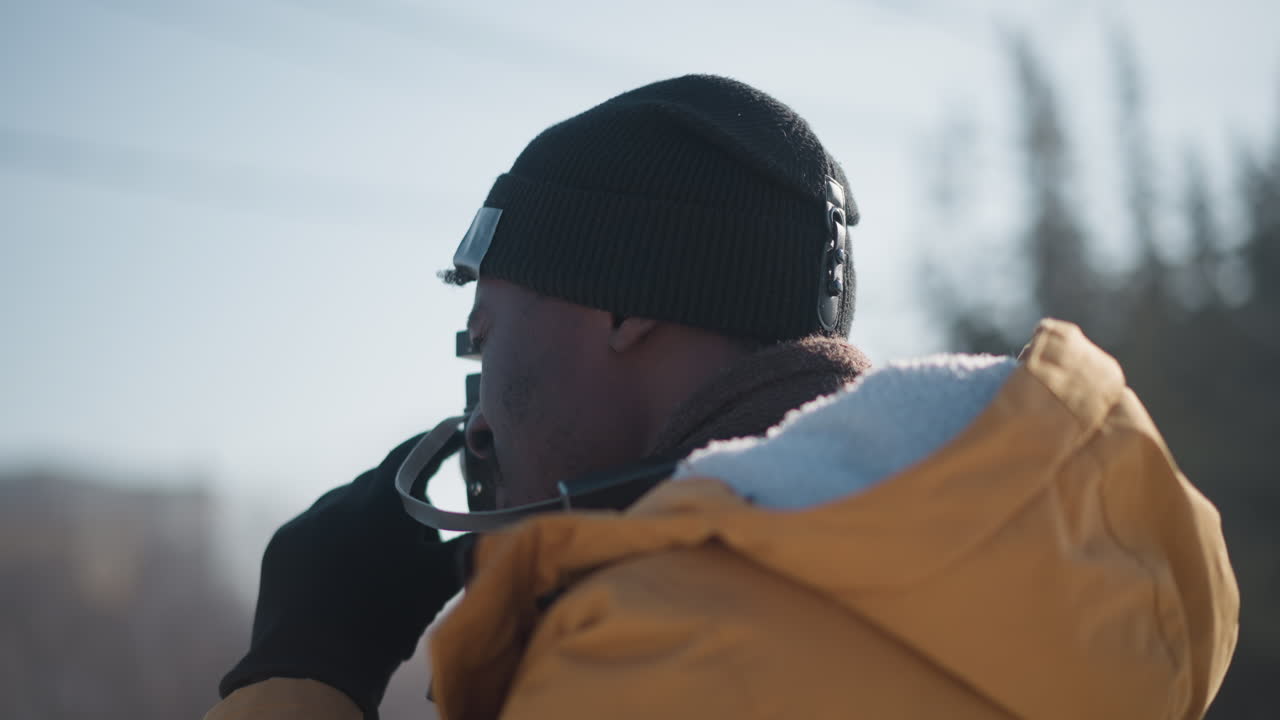 medium shot of cinematographer peering through viewfinder gloved hands adjusting focus dial under clear urban winter sky bright sunlight snowy pavement parked cars backdrop capturing urban scene