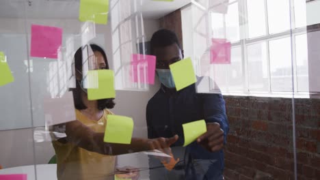 Diverse-male-and-female-business-colleagues-wearing-face-masks-putting-sticky-note-on-glass-wall