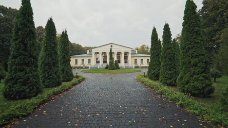 nasser bürgersteig, der zum großen leichten gebäude im park führt. schöne gasse mit grünen bäumen und einer schönen architektur im friedlichen landschaftsgarten im herbst.