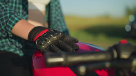 close-up of a gloved hand resting on the tank of a red motorcycle, with the rider s flannel shirt slightly lifted, revealing part of their waist