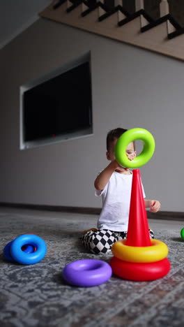 toddler boy in white t-shirt and checkered pants runs up to his toy. kid builds a pyramid at home.