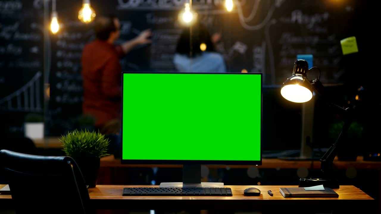 Personal Computer with Mock-up Green Screen Monitor Stands on the Office Desk, In the Background People Work on a Blackboard Wall in the Creative Office. Timelapse.