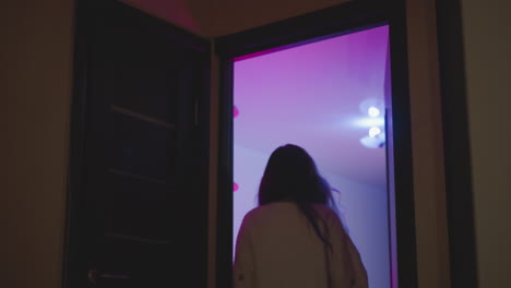 back view of woman in fluffy sweater walking through partially open door into neon tinted studio illuminated by overhead white bulb and bathed in pink purple ambient hue