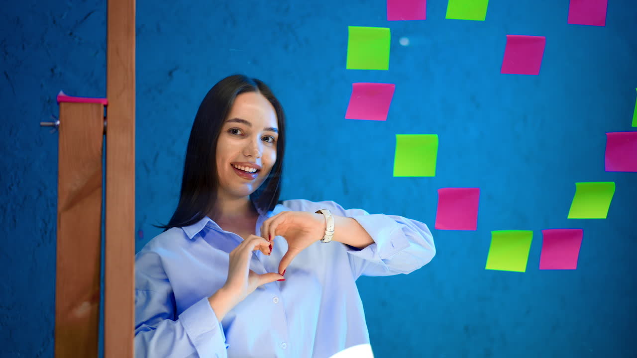 Woman creates heart with notes. A young woman joyfully arranges colorful sticky notes into a heart shape on a glass surface in a bright room