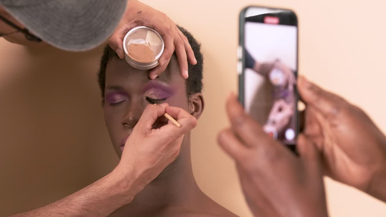 Makeup artist recording video on a mobile phone while doing makeup on a transgender person.