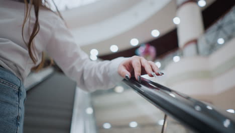 partial view of a lady with black polished nails gently tapping an escalator handrail while descending, she wears a white blouse and jeans, the blurred background captures a modern shopping mall