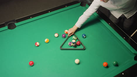 sports person in white shirt arranges colorful billiards using triangle rack on green pool table in dark room. focus, precision, and organization evident as balls are positioned for strategic gameplay