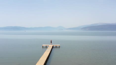 Aerial-shot-of-woman-enjoying-life-and-standing-on-a-beautiful-still-lake-with-mountains-in-the-horizon