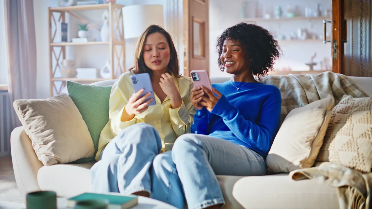 Female friends sharing phones sofa. Two young women browsing smartphones home.