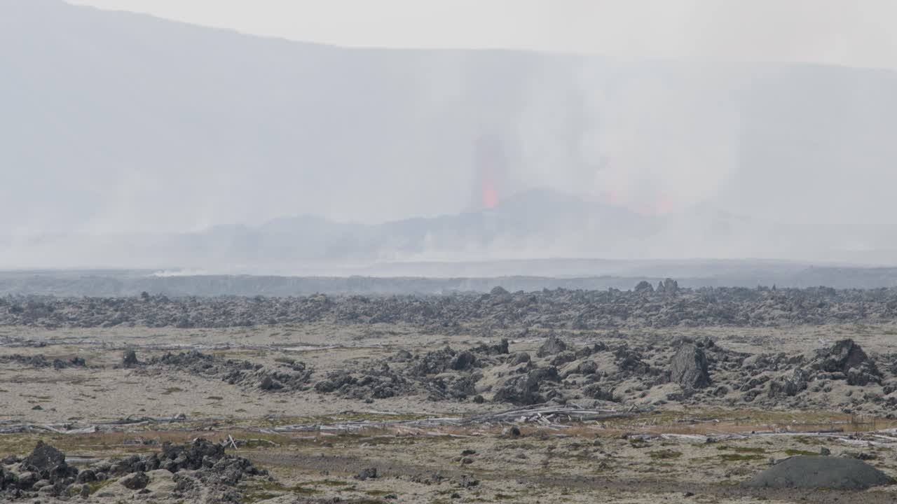 The camera tilts up from cars parking to reveal the erupting Grindavik volcano in Sundhnúkur crater, Iceland, visible in the distance.