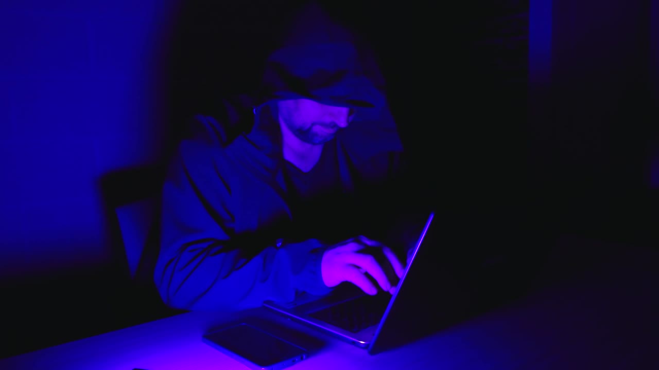 A man in a hooded jacket types on a laptop in a dark room illuminated with intense blue and purple lighting, evoking a cyber or hacker theme