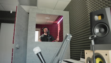 a man dressed in black stands in front of a studio microphone, wearing headphones as he moves his hands while singing in a red soundproofed room. the image also includes a speaker in the background