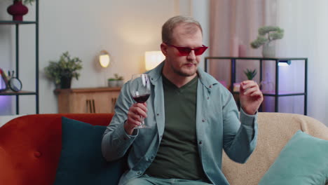 hombre adulto joven sonriente bebiendo una copa de vino rojo celebrando el aniversario de cumpleaños ganando la lotería en casa