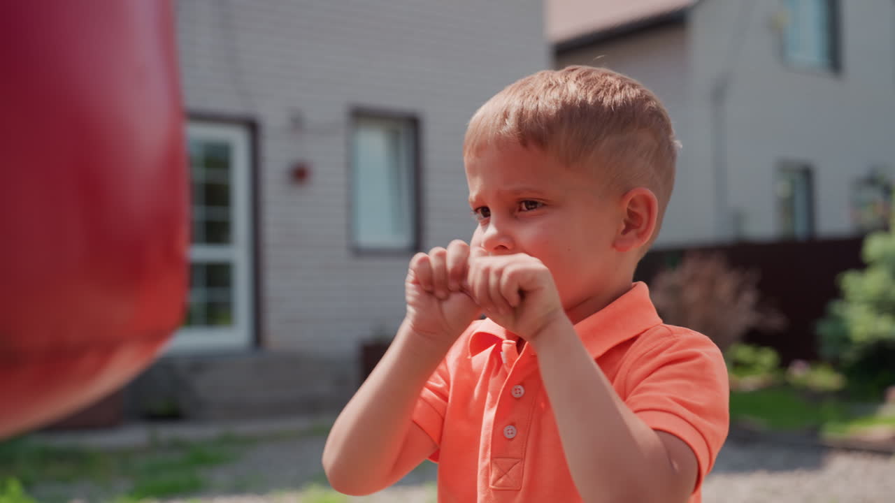 Child Strikes Red Bag Happily, Young Boy Enthusiastically Punches Bright Red Bag In Sunlight, Energetic Caucasian Boy Delivers Playful Punches To Colorful Bag Outdoors In Sunny Yard