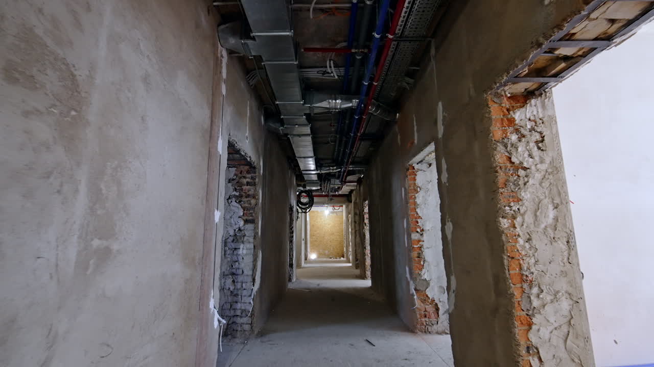 Movement by the long corridor being under reconstruction. Hall with grey unfinished walls and pipes on the ceiling.