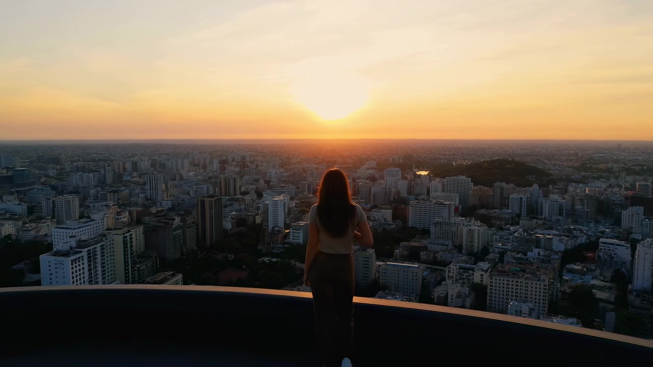 Vista traseira mostra figura solitária permanecendo perto do corrimão enquanto o enquadramento se alarga gradualmente, abrindo um amplo panorama urbano com edifícios agrupados, primeiro plano escuro e sol brilhante perto do horizonte.