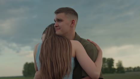 back view closeup of woman embracing man in open field at sunset, long hair flowing, eyes closed in gentle smile, affectionate bond under soft light and dramatic sky, intimate outdoor moment