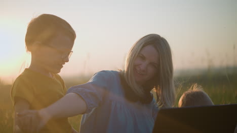 close-up of a woman in a blue gown sitting in a grassy field at sunset, playfully trying to stop a young boy dressed in yellow from distracting her while a little boy in white sits beside her