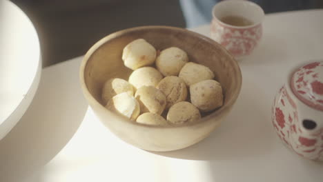 sunlight illuminates womans hand grabbing freshly baked chipa bread from wooden bowl, accompanied by a floral teapot and cup on a white table, creating a cozy home atmosphere footage