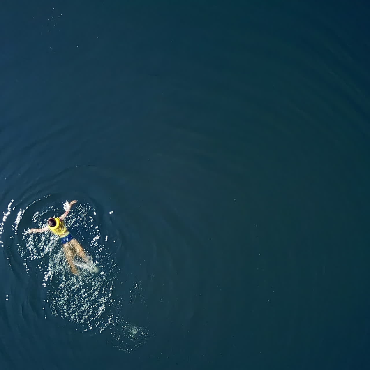 Little boy is swimming in a clear blue water in a sunny day in summer. Teenage boy wearing yellow life vest bathing alone on the water backgound. Aerial view