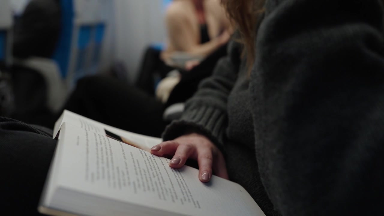Persona che legge un libro aperto con la mano appoggiata sulla pagina su un treno giapponese con passeggeri sullo sfondo