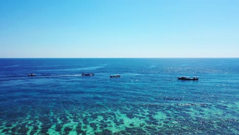 Boats-sailing-around-tropical-island's-shoreline-on-crystal-water-of-blue-turquoise-lagoon-with-beautiful-coral-reefs-underwater,-Jamaica