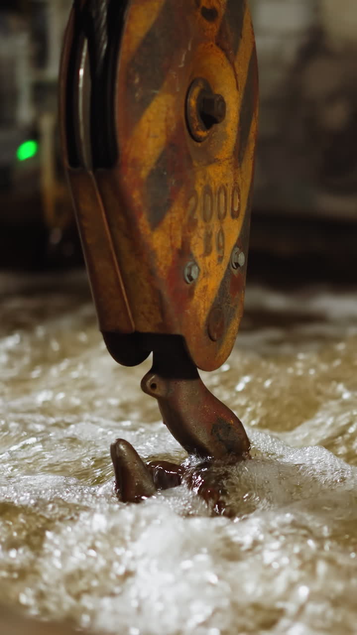 iluminado por una iluminación sutil, un gancho de grúa circula a través del agua burbujeante en un tanque de extinción industrial, causando una superficie de agua vaporosa y dinámica