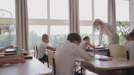 Student-in-protective-mask-studying-at-school-during-corona-virus-pandemic.-A-masked-teacher-explains-a-new-lesson-topic-to-masked-students.-children-write-with-pens-in-the-classroom