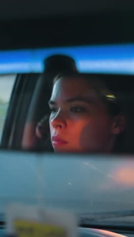 woman is reflected in rearview mirror while driving her car in the evening, with parked cars, and nearby buildings visible in the background under dim lighting in an urban road having a yellow mark