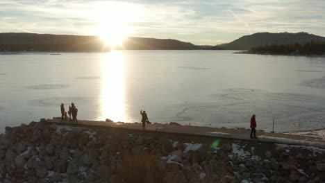 Panning-shot-while-tilting-down-of-people-silhouetted-on-pier-along-frozen-lake-at-sunset-hour