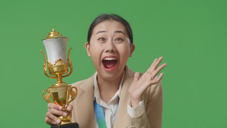 close up of asian business woman in a suit with a gold medal looking at a gold trophy in her hands and saying wow being happy winning as the first winner on green screen background in the studio