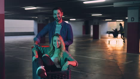 young caucasian man and woman looking at each other and smiling to the camera in a empty parking while woman is sitting in a trolley