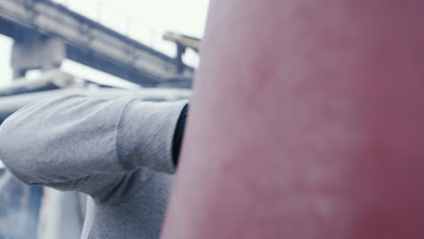 Close-up view of caucasian man in sportswear hitting a punching bag outdoors an abandoned factory on a cloudy morning