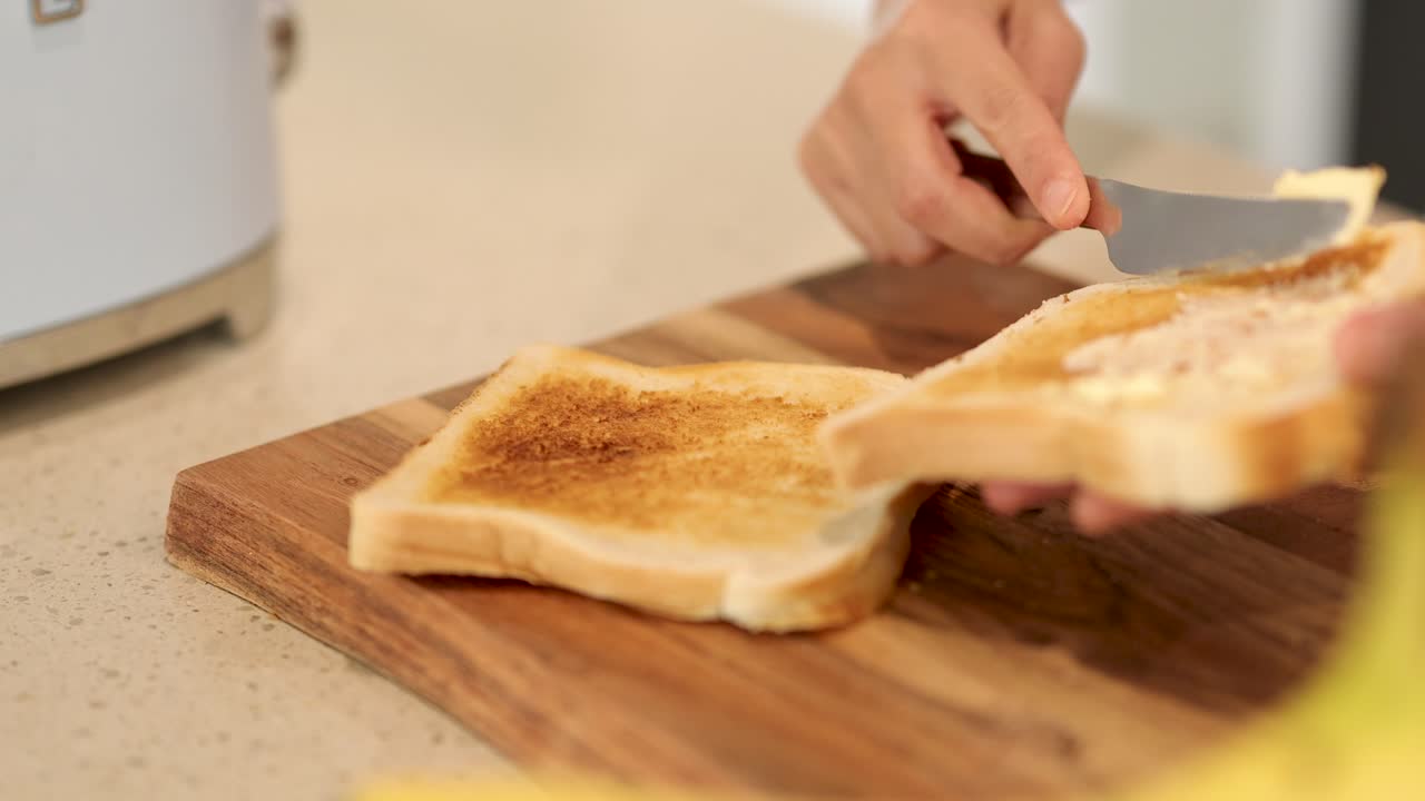 A person spreads butter on toasted bread using a knife on a wooden board in a well-lit kitchen