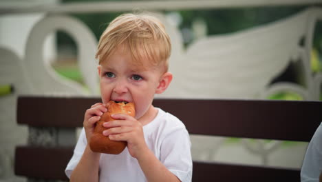 a young boy is seen seated and focused as he tries to take a larger bite of a sausage roll, there's a partial view of someone nearby in a white outfit