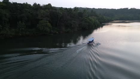 a tranquil drone view of a boat gliding smoothly along a goan river india, leaving soft ripples behind, surrounded by dense, lush green trees on both sides