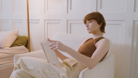 Vista-Lateral-De-Una-Chica-Sentada-En-Una-Silla-Leyendo-Un-Libro-En-El-Dormitorio