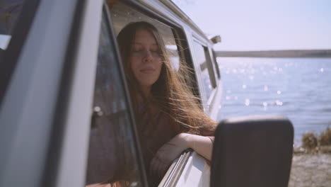 a young long haired red haired girl sitting in a caravan by a lake looks at the camera